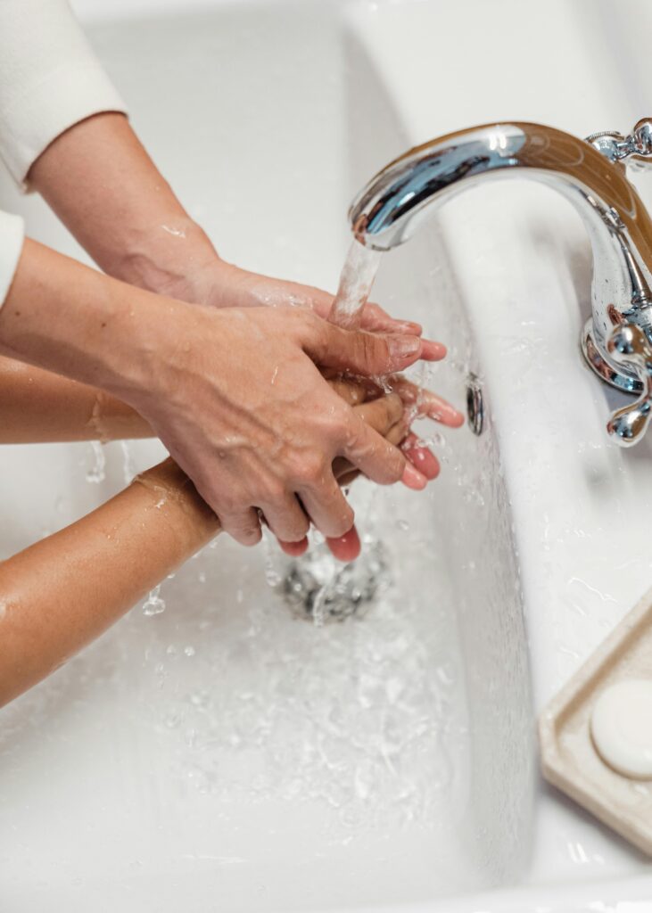 From above of crop unrecognizable mum washing hands of child with clear warm running water in white washbasin